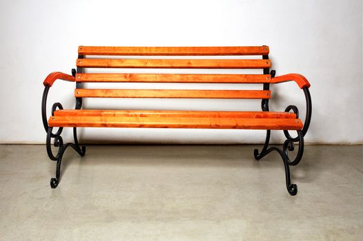 A beautiful wooden bench for rest with a metal forged base for landscaping walking areas in city parks against a white wall of a building and concrete floor, copy space.