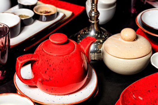 Red porcelain shiny teapot with black dots on a black table among other tea utensils, plates and handmade containers. Selective focus.