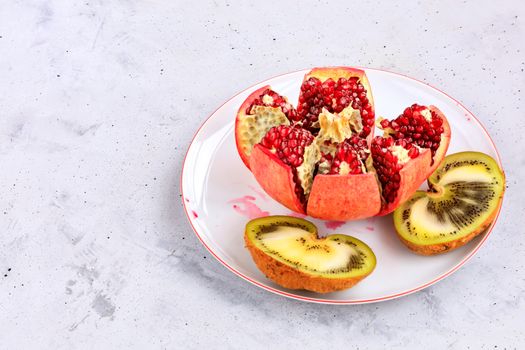 Ripe, red pomegranate fruit is divided into parts and a kiwi cut in half on a white porcelain plate, on a background of gray concrete, close-up, copy space.