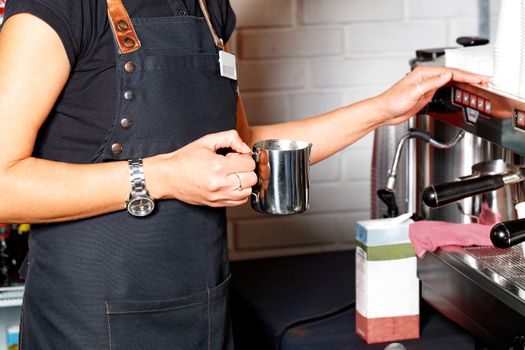The barista in a black apron holds in his hand a metal mug for making coffee latte on the background of a coffee machine and a packet of cream in blur.