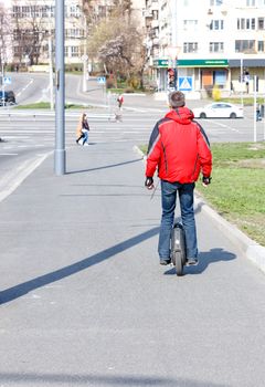 A man in a red jacket moves on an electric mono wheel along the sidewalk of a city street, a modern concept of freedom of movement.