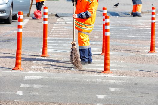 A janitor in a bright orange uniform sweeps the street and removes trash on the roadway and near the fence from the orange road columns.