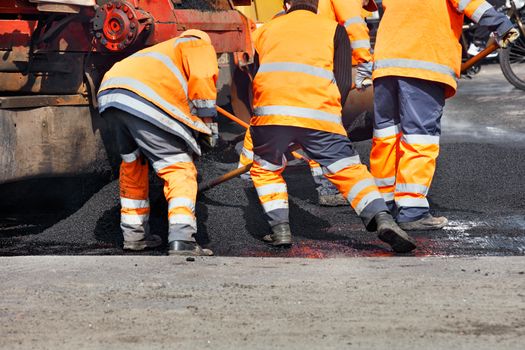 A team of road workers in an orange uniform updates part of the road with fresh asphalt and aligns it with shovels, to then compact it with a road roller, an image with copy space.