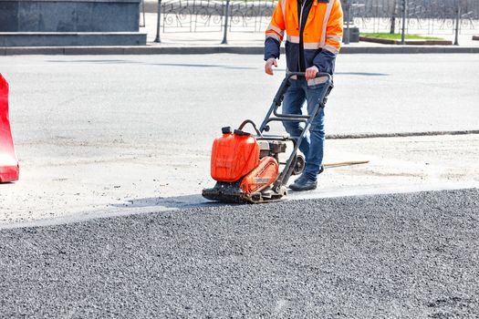 Worker road service use vibratory plate compactor compacting asphalt at road repair site.