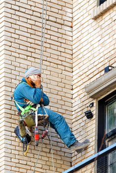 An industrial climber builder hangs on safety ropes on a brick wall of an old building with a set of various tools tied behind his back and receives phone instructions, a picture with copy space.