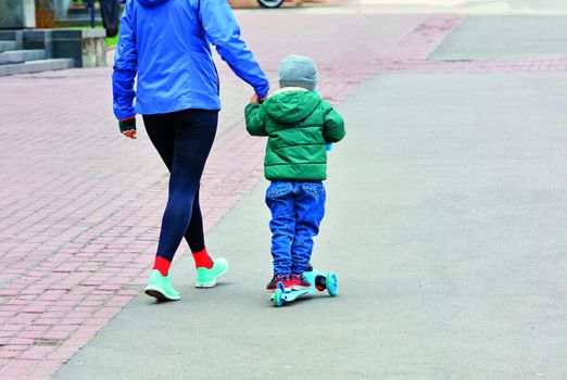 A child in funny baggy blue jeans and a green jacket on a children's scooter and his young mother in a sports blue jacket walk on the asphalt sidewalk of a city street.