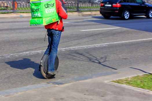 A young man from the Uber delivery service with a green backpack on his back rides an electric unicycle and delivers food. Courier on the electric monowheel delivering food. March 29, 2020.