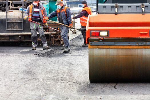 A working team of road workers in protective medical masks and overalls, using powerful road equipment and shovels, lay a new asphalt coating on the work site, image with copy space.