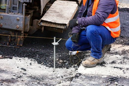 The worker sets the necessary level for the asphalting of the road with a tightly drawn cord against the background of the construction elements of a large paver.