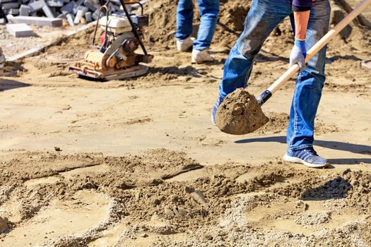 A worker levels the foundation with sand and a shovel under a wooden level for laying paving slabs against the background of the workplace and preparing for work, copy space.