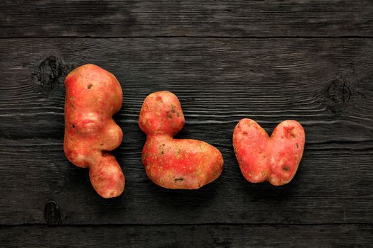 Ugly funny vegetables, heart-shaped potatoes and letter-shaped potatoes, black wooden background. The concept of grungy vegetables or food waste. Flat lay, copy space.