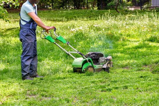 A utility worker in blue overalls looks after a green lawn with a petrol lawnmower.
