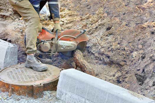 A worker using a portable concrete cutter and cutting a diamond blade cuts the powerful roots of a tree, which interfere with equipping the sewer hatch of the city collector.