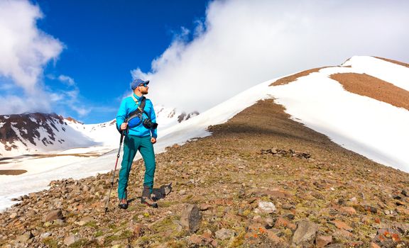 A male sports athletic photographer with a cane in his hand and a camera over his shoulder stands on a rocky slope of a snowy mountain and examines the top of a snowy mountain, planning a future hike.