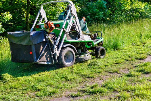A professional tractor lawn mower mows tall grass along a park grove, climbing up the slope and managed by a utility worker, copy space.