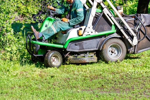 A service worker mows grass on a lawn with a tractor cross-country lawnmower on a clear sunny day, copy space.