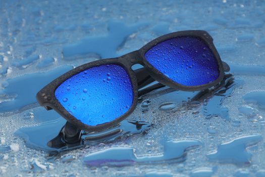 A pair of blue sunglasses with water drops on water reflection