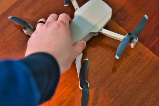 Hand holding drone with wooden deck visible in background. White man's hand grasping small flying drone preparing it to take off.