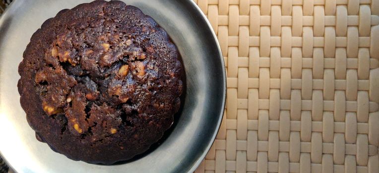 A close up of hot chocolate walnut muffin on a steel plate on a weave pattern table