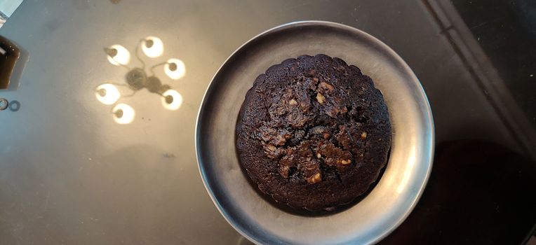 A chocolate walnut muffin on a steel plate against a reflective surface reflecting chandelier lights