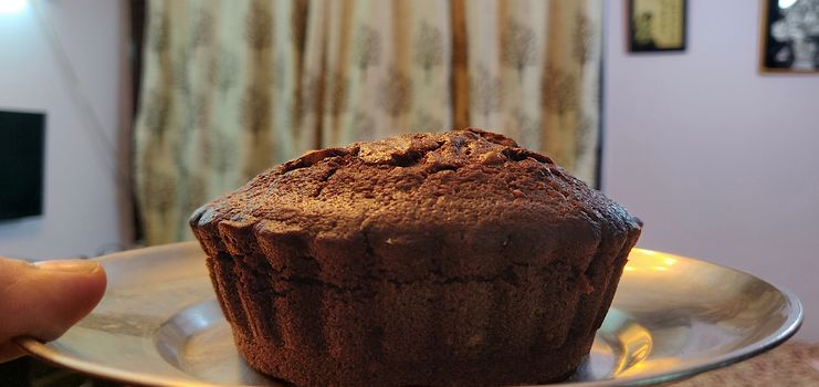 A close up of chocolate walnut muffin on a steel plate held up in the air by a hand against the wall of the house