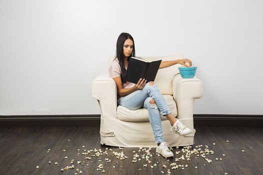 young woman sitting on a white couch, reading a hardcore book, eating popcorn from a blue bowl and making a mess