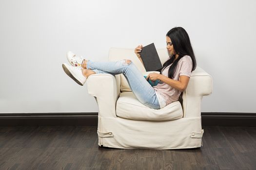 Young woman with her leg up on a white couch, eating from a blue bowl and reading a book