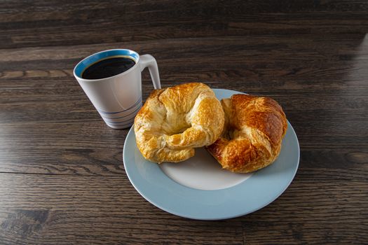 two croissants on a blue rimmed plate with a black coffee against a wood background