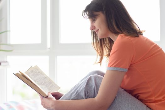 Girl at home by the window in the bedroom reads her favorite book