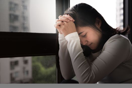 depressed women sitting near window and praying, alone, sadness, emotional concept