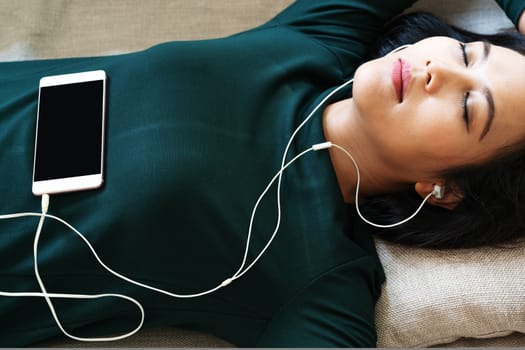 Over head view of beautiful young woman laying down on sofa using smartphone to listen the music
