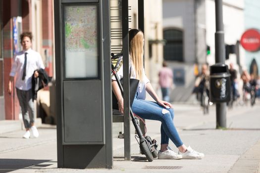 Casual caucasian teenager commuter with modern foldable urban electric scooter sitting on a bus stop bench waiting for metro city bus. Urban mobility concept.