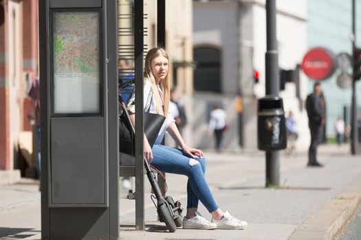 Casual caucasian teenager commuter with modern foldable urban electric scooter sitting on a bus stop bench waiting for metro city bus. Urban mobility concept.