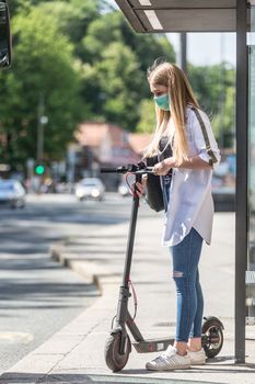 Casual caucasian teenager commuter waering protective face mask against spreading of corona virus with modern foldable urban electric scooter waiting for metro city bus. Urban mobility concept.