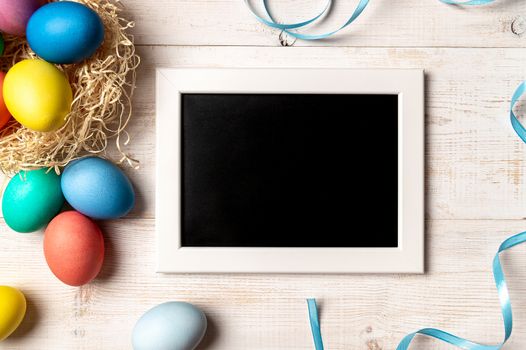 Easter concept. Colorful eggs on white wooden background with empty chalkboard. Copy space for greetings, text or design. Top down view or flat lay