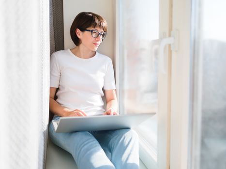 Woman remotely from home. She sits on window sill with laptop on knees. Lockdown quarantine because of coronavirus COVID19. Self isolation at home.