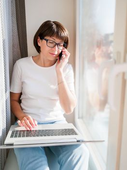 Woman works remotely from home. She sits on window sill with laptop on knees. Lockdown quarantine because of coronavirus COVID19. Self isolation at home.