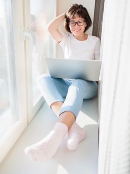 Smiling woman works remotely from home. She sits on window sill with laptop on knees. Lockdown quarantine because of coronavirus COVID19. Self isolation at home.