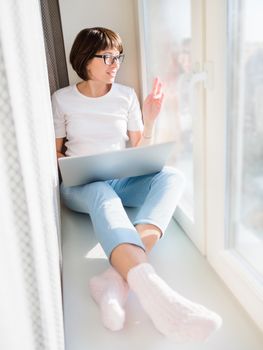 Smiling woman works remotely from home. She sits on window sill with laptop on knees. Lockdown quarantine because of coronavirus COVID19. Self isolation at home.