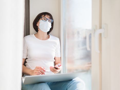 Woman in medical protective mask works remotely from home. She sits on window sill with smartphone and laptop on knees. Lockdown quarantine because of coronavirus COVID19.