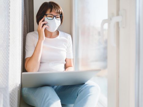 Woman in medical protective mask with smartphone works remotely from home. She sits on window sill with laptop on knees. Lockdown quarantine because of coronavirus COVID19. Self isolation.