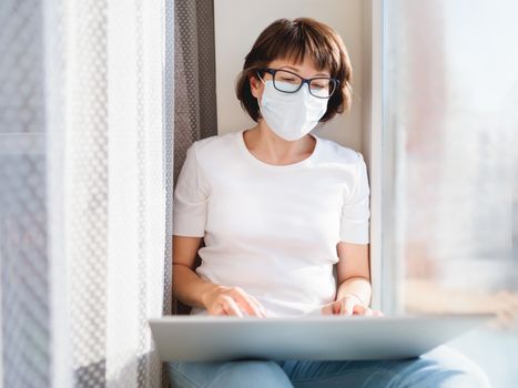 Woman in medical mask works remotely from home. She sits on window sill with laptop on knees. Lockdown quarantine because of coronavirus COVID19. Self isolation at home.