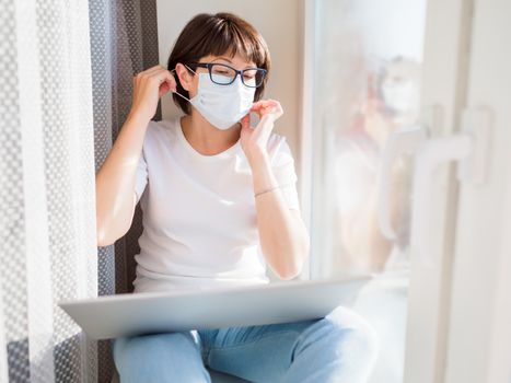 Woman in medical mask works remotely from home. She sits on window sill with laptop on knees. Lockdown quarantine because of coronavirus COVID19. Self isolation at home.