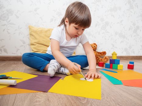 Toddler boy learns to cut colored paper with scissors. Kid sits on floor in kids room with toy blocks and teddy bear. Educational classes for children. Developing feeling sensations and fine motor skills at home.