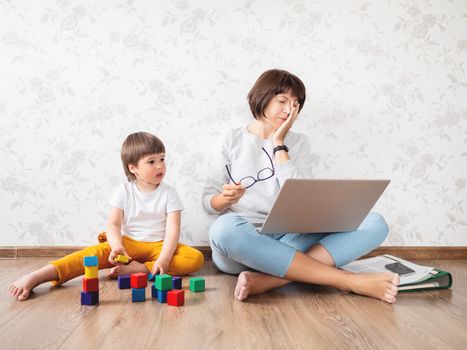Mom and son argue at home quarantine because of coronavirus COVID19. Tired mother works remotely with laptop, son plays with toy blocks. Self isolation at home.