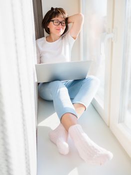 Smiling woman works remotely from home. She sits on window sill with laptop on knees. Lockdown quarantine because of coronavirus COVID19. Self isolation at home.