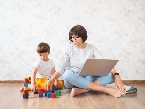 Mom and son argue at home quarantine because of coronavirus COVID19. Mother works remotely with laptop, son plays with toy blocks. Self isolation at home.
