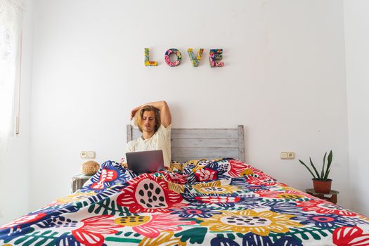 Young man with medium long blond hair, with a beard and mustache working with the computer from home during quarantine, from bed. Concept adapt to the new reality