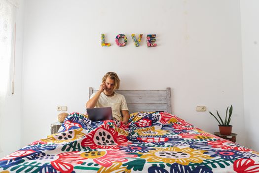 Young man with medium long blonde hair, with a beard and mustache working on the computer from home during quarantine, from bed. Calling by phone. Concept adapted to the new reality.