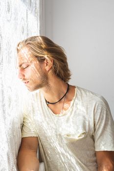 Young man with medium long blonde hair frustrated at not being able to leave home. Loneliness and silence concept. Sun rays coming into the house through the window.